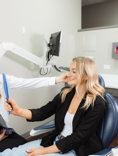 A woman with long blonde hair, wearing a black blazer, smiles while holding a blue mirror in a dental office chair. A person in a white coat gently touches her face, and a dental monitor is visible in the background.