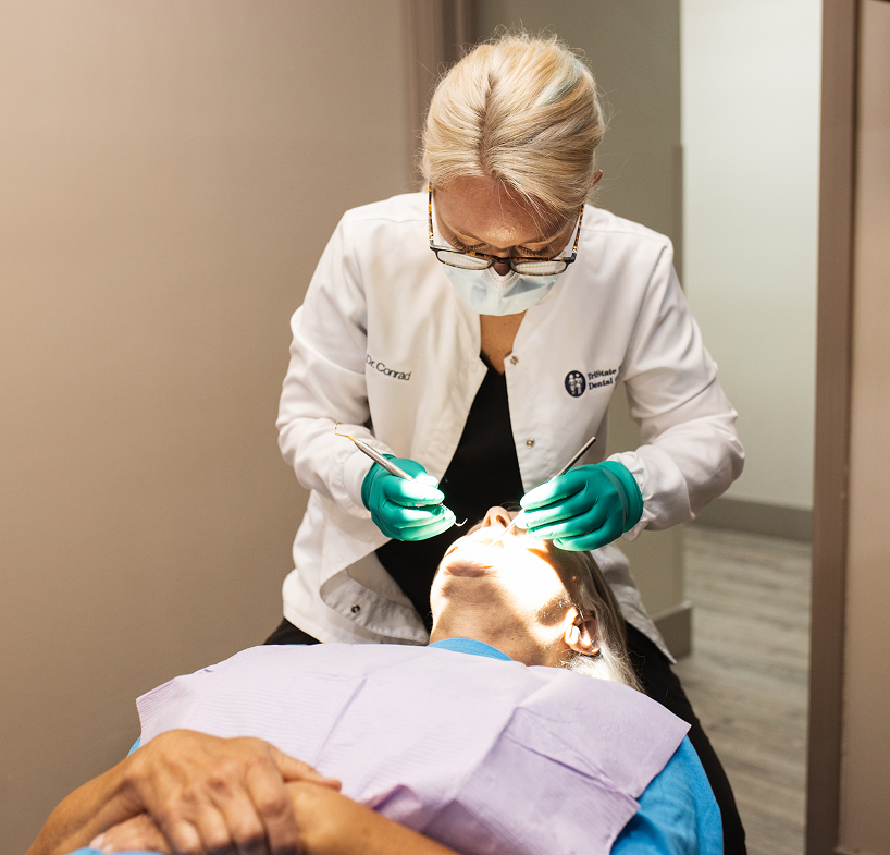 A masked dentist in a white coat and green gloves examines a patient lying back in a dental chair with a purple bib.