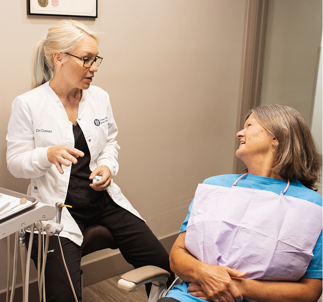 Dentist in a white coat speaks with a smiling patient in a blue shirt and dental bib during a calm, friendly consultation.